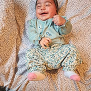 Inaya is registered to the contest to win money with this photo: baby, child, smiling, headband, floral_pants, patterned_bedspread, happy, lying_down, infant, cute, clothing, socks, playful, indoors, young_child, person, joyful, cheerful, hands, face