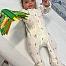 baby, child, expression, face, floor, footwear, hand, indoor, infant, lying_down, medical_table, onesie, patterned_clothing, person, playful_wall_art, raccoon_design, soft_book, toy, vegetables, white_background