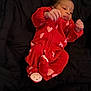baby, infant, red_onesie, hearts, black_blanket, lying_down, tongue_out, cute, sleepwear, newborn, small_hands, footed_pajamas, playful, child, indoors, cozy, soft_fabric, person, resting, adorable