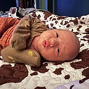 Presley is registered to the contest to win money with this photo: baby, newborn, infant, blanket, cow_pattern, bed, quilt, brown, white, clothing, mittens, face, head, person, lying_down, indoors, cute, expression, soft, warm