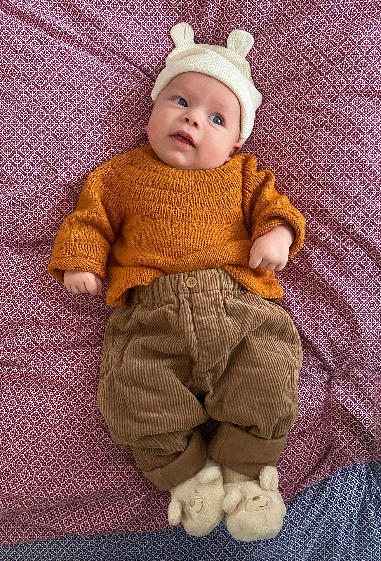 Félix participe au concours pour gagner de l'argent avec cette photo : baby, infant, child, newborn, hat, bear_hat, knitted_sweater, mustard_sweater, corduroy_pants, patterned_sheet, bedding, slippers, plush_slippers, lying_down, portrait, eyes_open, cute, cozy, hand, curious_expression