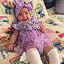 baby, infant, smiling, purple_dress, headband, bow, bed, bedding, cartoon_characters, stitch, cute, happy, white_socks, toddler, patterned_bedspread, child, indoor, portrait, soft_toys, colorful