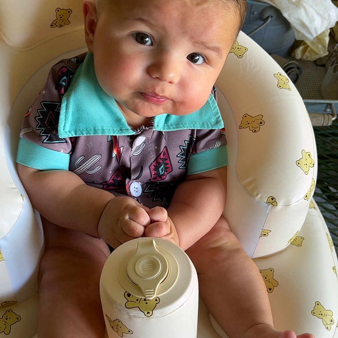 Lucas is registered to the contest to win money with this photo: baby, child, clothing, colorful_shirt, container, curious_expression, cushion, cute, face, foot, hand, indoors, infant, looking_at_camera, person, portrait, seat, sitting, skin, teddy_bear_print