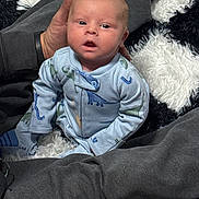 Noah is registered to the contest to win money with this photo: baby, infant, dinosaur_onesie, hand, adult, rug, black_and_white, shaggy_rug, clothing, person, cute, indoor, portrait, sitting, looking_up, soft_texture, comfort, child, young, cozy