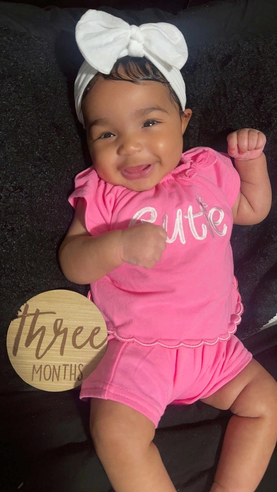 Novie joined the competition — help win amazing prizes! baby, infant, smile, pink_clothing, headband, bow, cute, happy, lying_down, milestone, wooden_plaque, three_months, fingers, cheeks, black_blanket, cute_expression, portrait, child, young_baby, celebration