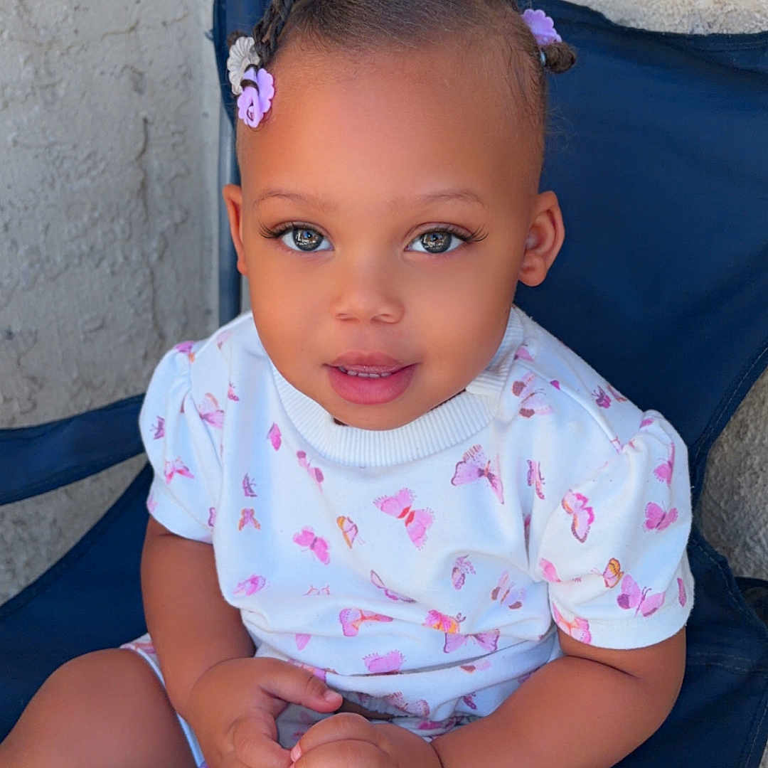 Ruby is registered to the contest to win money with this photo: child, toddler, baby, portrait, sitting, outdoor, chair, blue_chair, butterfly_print, floral_clothing, braided_hair, hair_bows, smiling, face, eyes, hands, legs, textured_wall, patio, photography