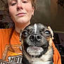 person, dog, small_dog, close_up, selfie, earrings, orange_shirt, harley_davidson, indoor, smile, pet, collar, brown_hair, face, happy, cute, animal, portrait, young_adult, casual
