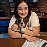 woman, person, dog, puppy, boxer, pet, portrait, smiling, hugging, restaurant, pub, indoor, booth, table, brick_wall, chalkboard, pendant_light, lottery_ticket, lighter, cozy