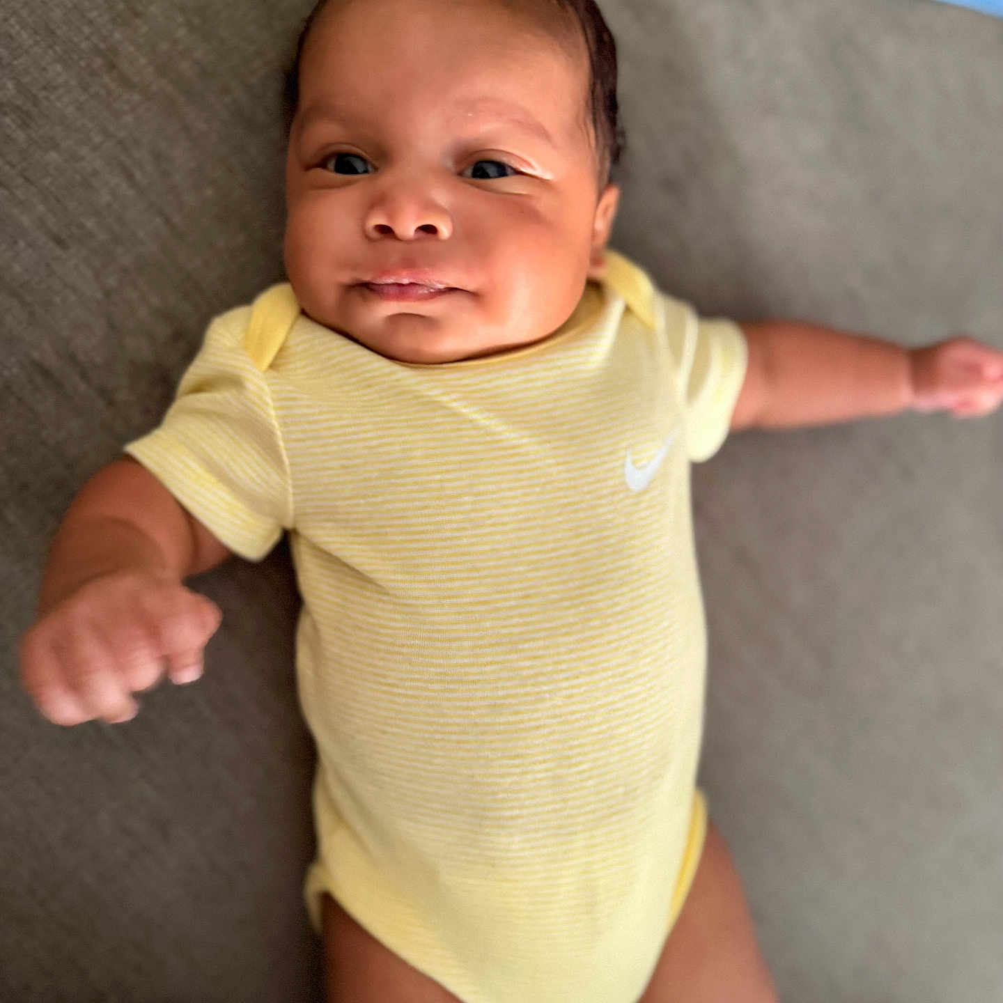 Ajayveon is registered to the contest to win money with this photo: arm, baby, blanket, child, cute, expression, eyes, face, hand, head, indoor, infant, lying_down, newborn, people, portrait, skin, smile, soft_surface, yellow_onesie