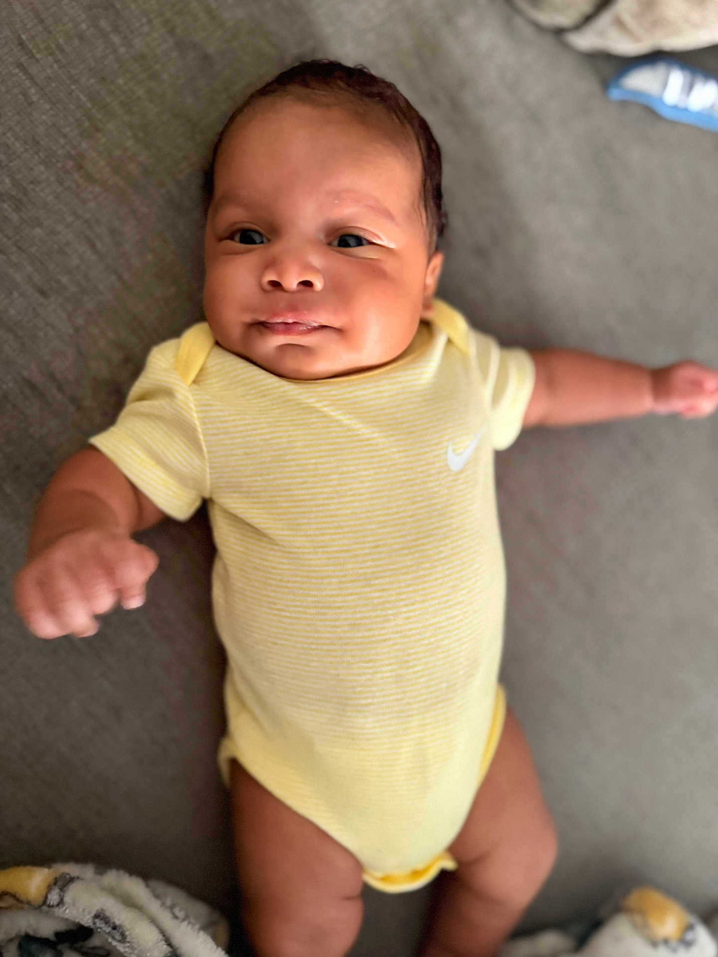 Ajayveon is registered to the contest to win money with this photo: baby, infant, yellow_onesie, lying_down, face, skin, arm, hand, blanket, soft_surface, expression, cute, child, newborn, people, portrait, indoor, head, eyes, smile