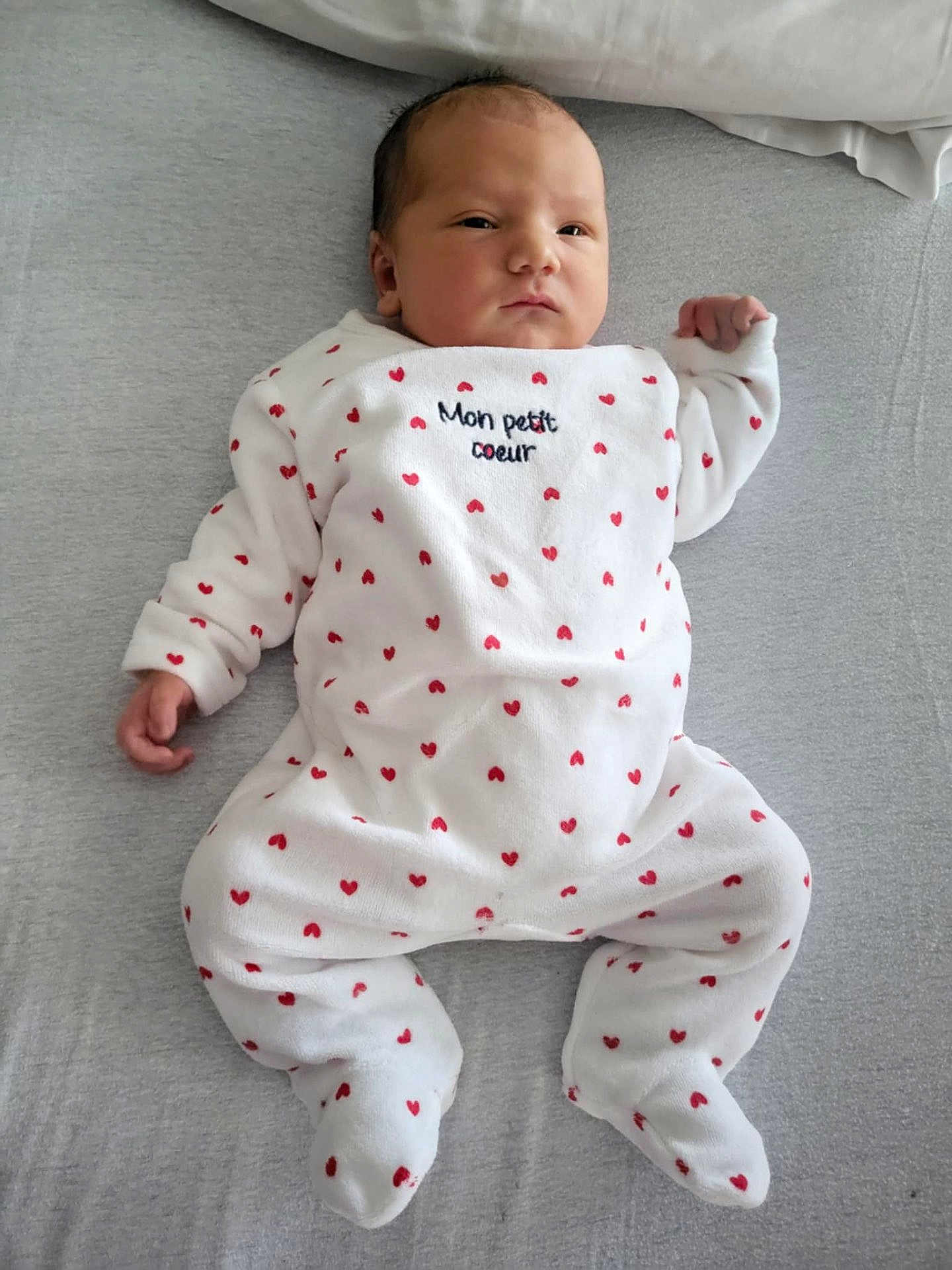 Nino participe au concours pour gagner de l'argent avec cette photo : baby, infant, onesie, white_clothing, red_hearts, lying_down, soft_surface, face, cute, child, newborn, person, sleepwear, bed, expression, calm, small_hands, footed_pajamas, indoors, portrait