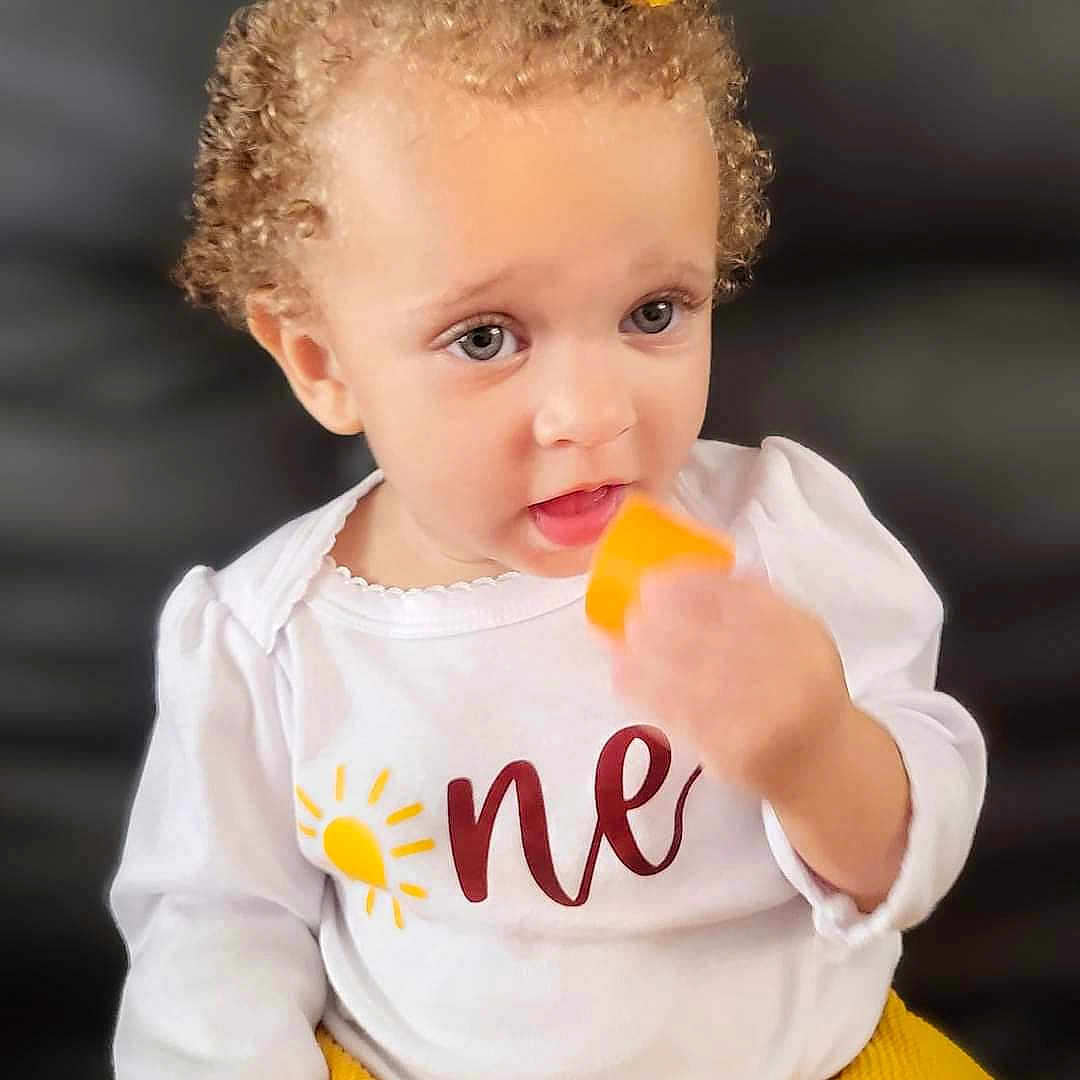 Stella Rose joined the competition — help win amazing prizes! baby, biting, blonde, bodypart, clothing, curlyhair, eating, face, finger, food, hair, hand, happy, head, person, photography, portrait, sitting, sleeve, smile