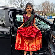 Mokshita is registered to the contest to win money with this photo: child, girl, traditional_clothing, red_skirt, gold_pattern, barefoot, car_door, black_car, parking_lot, jewelry, bracelet, outdoor, overcast_sky, tree, pose, portrait, young_person, cultural_dress, standing, vehicle