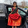 child, girl, traditional_clothing, red_skirt, gold_pattern, barefoot, car_door, black_car, parking_lot, jewelry, bracelet, outdoor, overcast_sky, tree, pose, portrait, young_person, cultural_dress, standing, vehicle