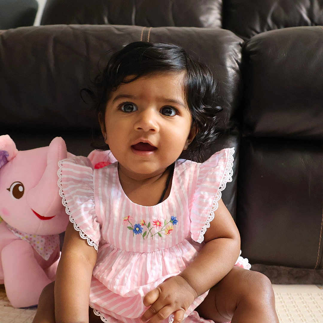 Sitara joined the competition — help win amazing prizes! baby, child, curly_hair, cute, decor, elephant_toy, floor, happy, home, indoor, leather_couch, person, pink_dress, playful, portrait, seated, smiling, soft_toy, stuffed_animal, young_child