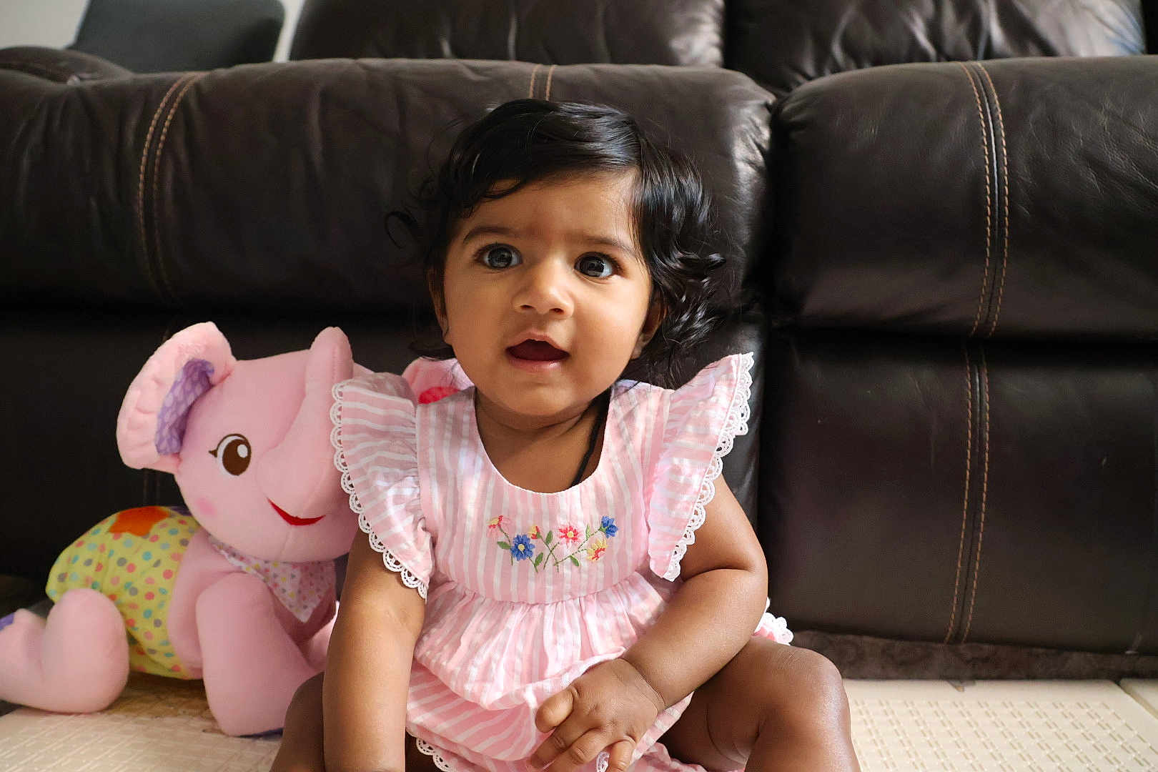 Sitara joined the competition — help win amazing prizes! baby, child, pink_dress, stuffed_animal, elephant_toy, indoor, leather_couch, curly_hair, floor, cute, portrait, smiling, seated, young_child, playful, soft_toy, home, person, happy, decor