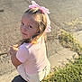child, girl, pink_bow, curly_hair, sidewalk, sunlight, outdoor, grass, crouching, smile, portrait, casual_clothing, young, daylight, nature, happy, face, person, cute, fashion
