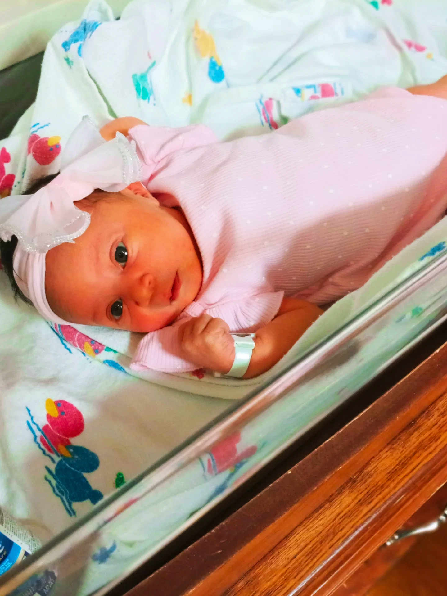 Nova is registered to the contest to win money with this photo: newborn, baby, infant, pink_outfit, headband, bow, hospital_bassinet, blanket, duck_pattern, curious, wide_eyes, lying_down, hospital, wooden_furniture, soft_fabric, cute, small_hand, wristband, portrait, indoors