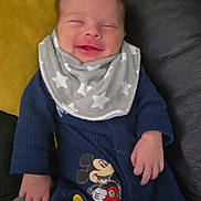 Luke joined the competition — help win amazing prizes! baby, smiling, infant, bib, mickey_mouse, blue_outfit, couch, leather, yellow_cushion, cozy, cute, child, happy, small_hands, relaxed, indoors, portrait, human, sitting, soft_texture