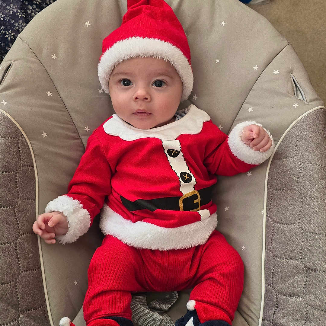 Luke is registered to the contest to win money with this photo: baby, baby_seat, carpet, child, christmas_hat, cute, festive, gift_bags, holiday_decor, holiday_season, indoor, infant, penguin_slippers, person, red_clothing, santa_outfit, small_child, smiling, soft_toys, winter_clothing