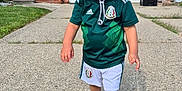 Diego joined the competition — help win amazing prizes! toddler, child, smiling, hat, soccer_jersey, shorts, shoes, sidewalk, driveway, house, grass, outdoor, daylight, person, happy, walking, concrete, casual_clothing, green, black_shoes