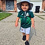 toddler, child, smiling, hat, soccer_jersey, shorts, shoes, sidewalk, driveway, house, grass, outdoor, daylight, person, happy, walking, concrete, casual_clothing, green, black_shoes