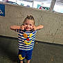 child, kid, playful, parking_garage, concrete_wall, striped_shirt, shorts, fruit_patches, white_sandals, silly_face, cheeks, urban, outdoor, standing, expression, casual_clothing, hair_tied, daylight, person, smile
