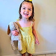 Ellie is registered to the contest to win money with this photo: child, girl, smiling, yellow_dress, hair_bow, plush_toy, bunny, barefoot, standing, indoor, floor, wall, happy, cute, portrait, young_child, toy, clothing, person, playful