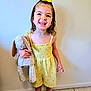 child, girl, smiling, yellow_dress, hair_bow, plush_toy, bunny, barefoot, standing, indoor, floor, wall, happy, cute, portrait, young_child, toy, clothing, person, playful