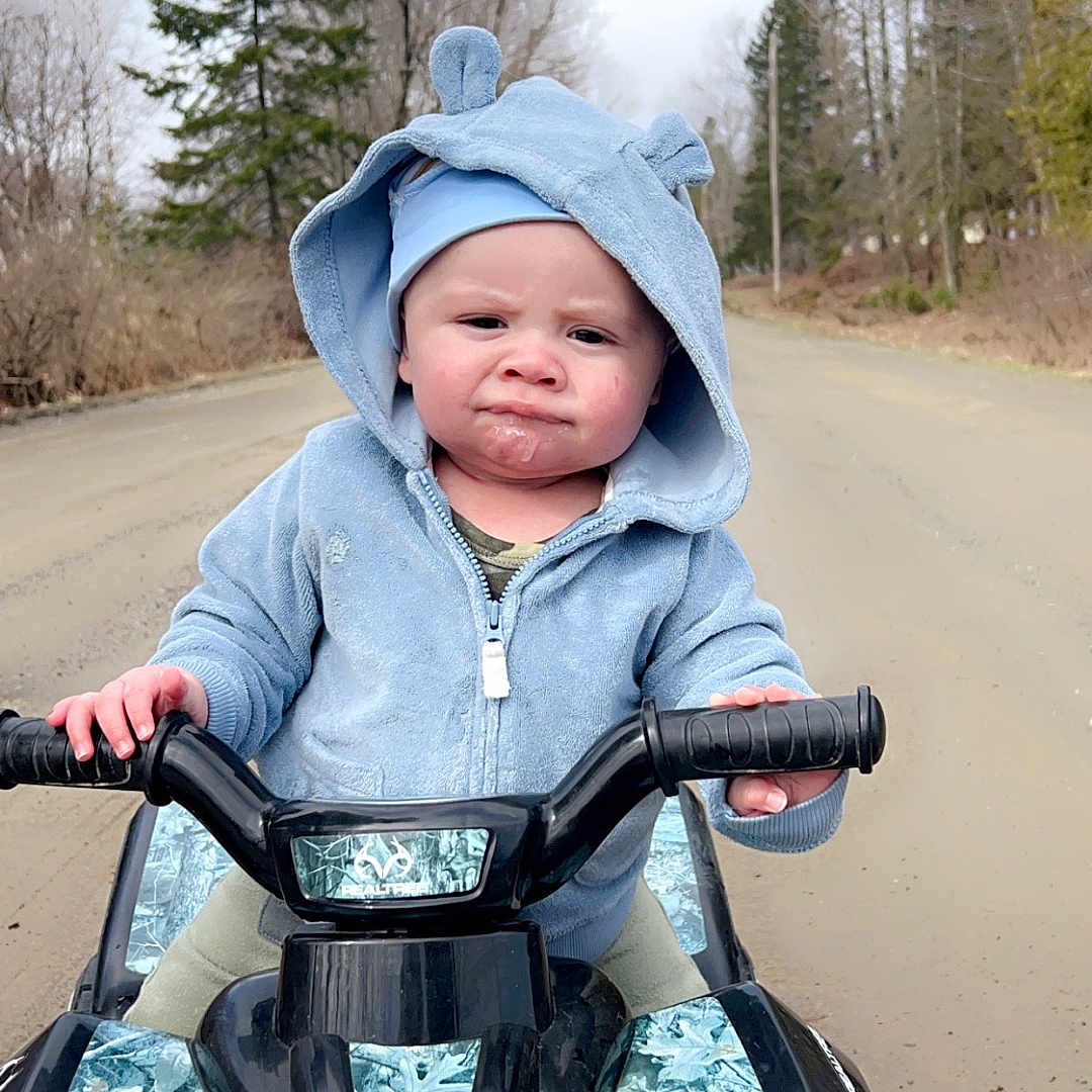 Cyrus is registered to the contest to win money with this photo: baby, clothing, coat, face, grass, head, hood, hoodie, jacket, knitwear, motorcycle, person, photography, plant, portrait, sweater, sweatshirt, transportation, tree, vegetation