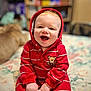 baby, child, red_onesie, hood, smiling, happy, face, hands, bed, blanket, floral_pattern, indoors, cute, infant, portrait, sitting, toddler, soft_focus, clothing, teddy_bear_patch