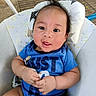 baby, child, infant, portrait, smiling, happy, sitting, blue_shirt, clothing, face, eyes, hair, hands, arms, cushioned_seat, pillow, outdoor, patio, daylight, close_up