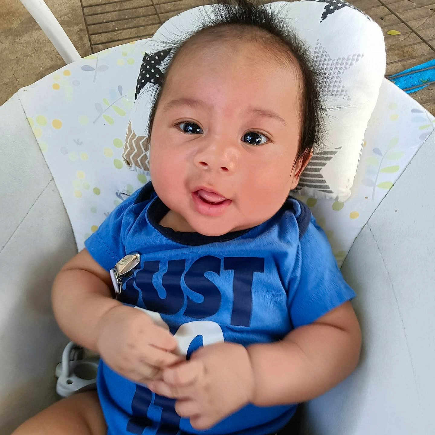 Kingston is registered to the contest to win money with this photo: baby, child, infant, portrait, smiling, happy, sitting, blue_shirt, clothing, face, eyes, hair, hands, arms, cushioned_seat, pillow, outdoor, patio, daylight, close_up