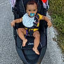 baby, pacifier, stroller, jeep, pavement, grass, yellow_clothing, bib, outdoor, child, infant, seatbelt, foot, hand, pathway, nature, curly_hair, sunlight, black_stroller, relaxed