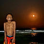 child, boy, smiling, beach, night, ocean, waves, moon, reflection, swim_trunks, bare_chest, water, outdoor, shore, happiness, tropical, nature, youth, vacation, fun