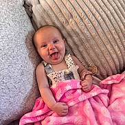 Emerson Rae is registered to the contest to win money with this photo: baby, blanket, child, clothing, couch, cozy, cute, happy, indoor, infant, person, pillow, pink, playful, portrait, relaxed, sitting, smiling, soft_texture, tongue_out
