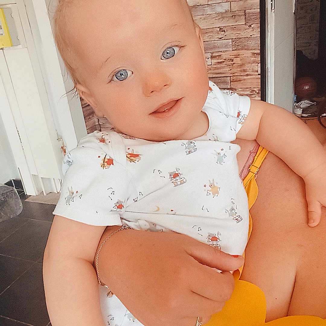 Morange Lyzeo participe au concours pour gagner de l'argent avec cette photo : adult, arm, baby, blue_eyes, bright, casual_clothing, child, cute, floor_tiles, hand, holding, home, indoor, infant, person, photograph, portrait, smiling, white_onesie, wooden_wall