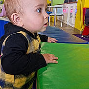Ethan participe au concours pour gagner de l'argent avec cette photo : background, black_clothing, chair, child, colorful, curious, face, floor, furniture, green_surface, hand, indoor, plaid_vest, play_area, spiky_hair, standing, table, toddler, wide_eyes, yellow_wall