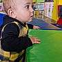 background, black_clothing, chair, child, colorful, curious, face, floor, furniture, green_surface, hand, indoor, plaid_vest, play_area, spiky_hair, standing, table, toddler, wide_eyes, yellow_wall