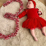 Ambre a rejoint le concours — aidez-le/la à gagner de superbes lots ! baby, red_dress, headband, tinsel, number_three, fluffy_rug, wooden_floor, infant, festive, holiday, cute, lying_down, celebration, child, indoors, soft_texture, decor, portrait, smiling, chubby_cheeks