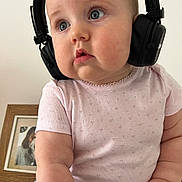 Mira participe au concours pour gagner de l'argent avec cette photo : baby, headphones, child, pink_shirt, blue_pants, floral_pattern, chubby_cheeks, curious_expression, indoor, portrait, close_up, person, cute, seated, hand, face, skin, young_child, clothing, home