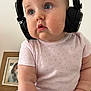 baby, headphones, child, pink_shirt, blue_pants, floral_pattern, chubby_cheeks, curious_expression, indoor, portrait, close_up, person, cute, seated, hand, face, skin, young_child, clothing, home