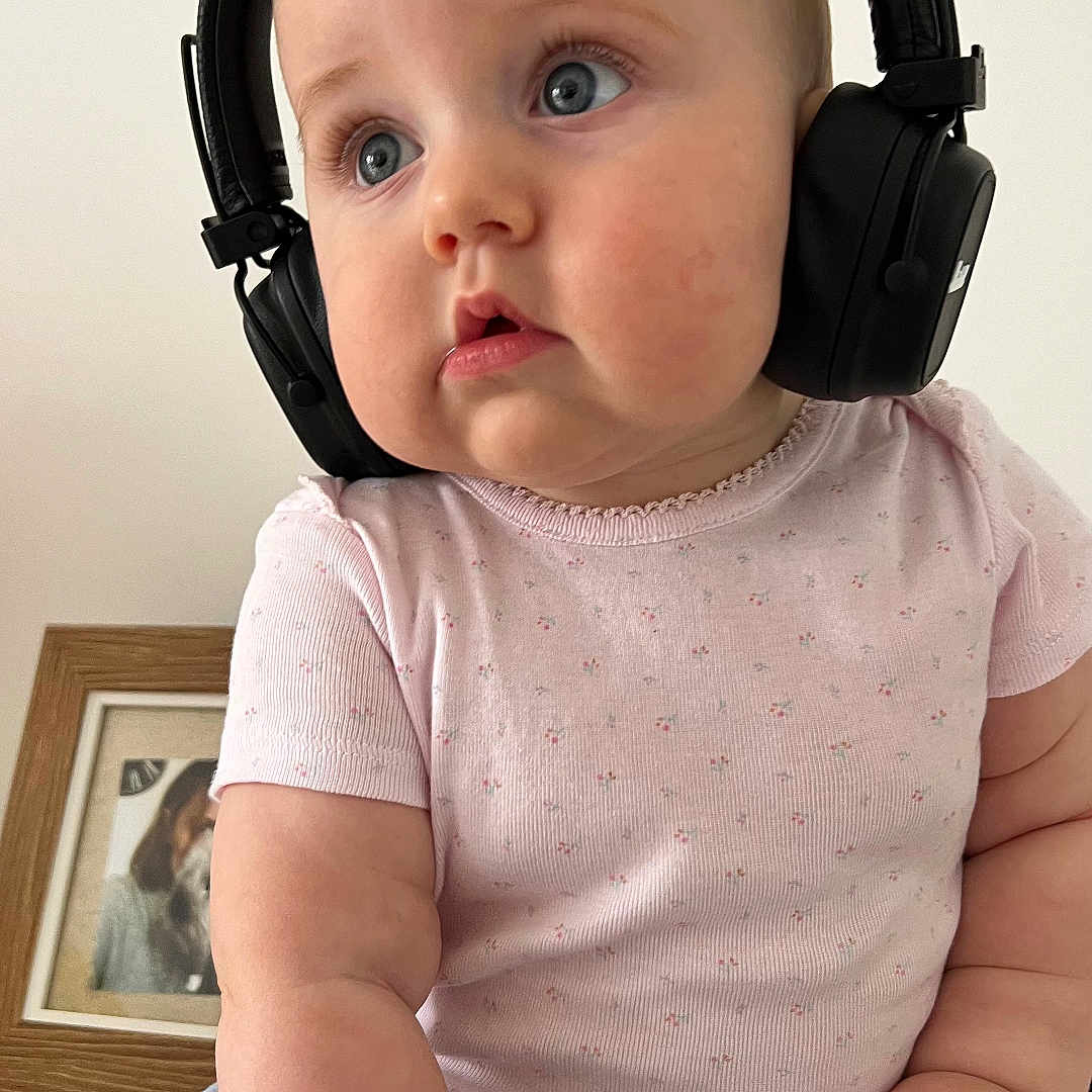 Mira participe au concours pour gagner de l'argent avec cette photo : baby, blue_pants, child, chubby_cheeks, close_up, clothing, curious_expression, cute, face, floral_pattern, hand, headphones, home, indoor, person, pink_shirt, portrait, seated, skin, young_child