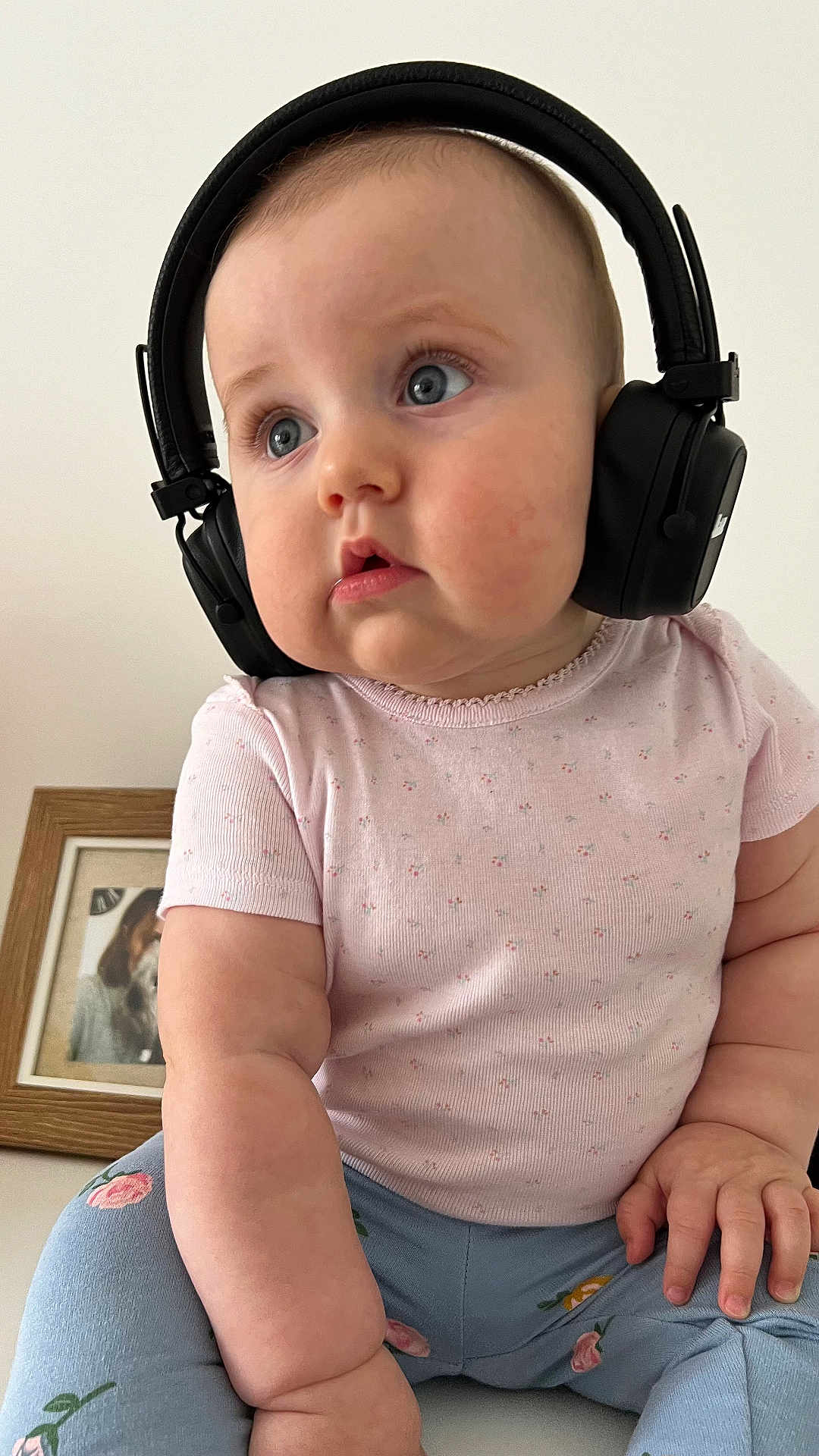 Mira participe au concours pour gagner de l'argent avec cette photo : baby, headphones, child, pink_shirt, blue_pants, floral_pattern, chubby_cheeks, curious_expression, indoor, portrait, close_up, person, cute, seated, hand, face, skin, young_child, clothing, home