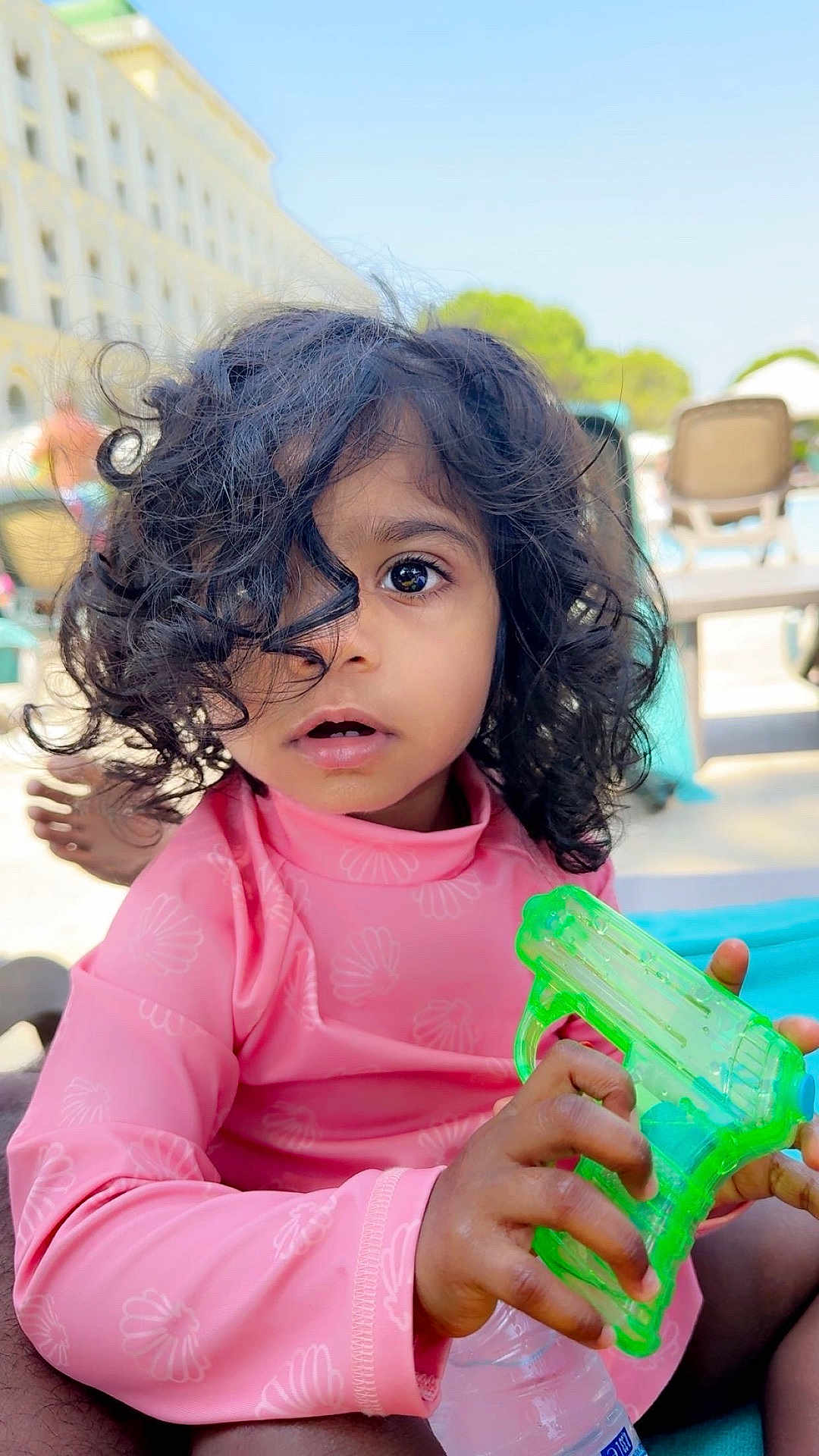 Léa a rejoint le concours — aidez-le/la à gagner de superbes lots ! child, toddler, curly_hair, pink_clothing, water_gun, outdoor, sunny, poolside, lounge_chair, expression, portrait, hand, summer, playful, toy, young, casual, daylight, skin, person
