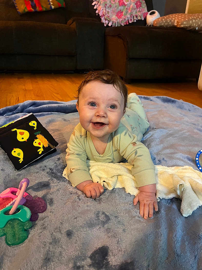 Paisley is registered to the contest to win money with this photo: baby, infant, tummy_time, smile, blanket, soft_book, toy, teether, pajamas, towel, drool, hands, face, eyes, living_room, couch, hardwood_floor, playmat, indoor, happy