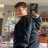 person, dog, chihuahua, pet, portrait, smiling, glasses, hoodie, black_clothing, standing, indoor, dining_room, table, tablecloth, bookshelf, speaker, framed_pictures, home, small_dog, playful