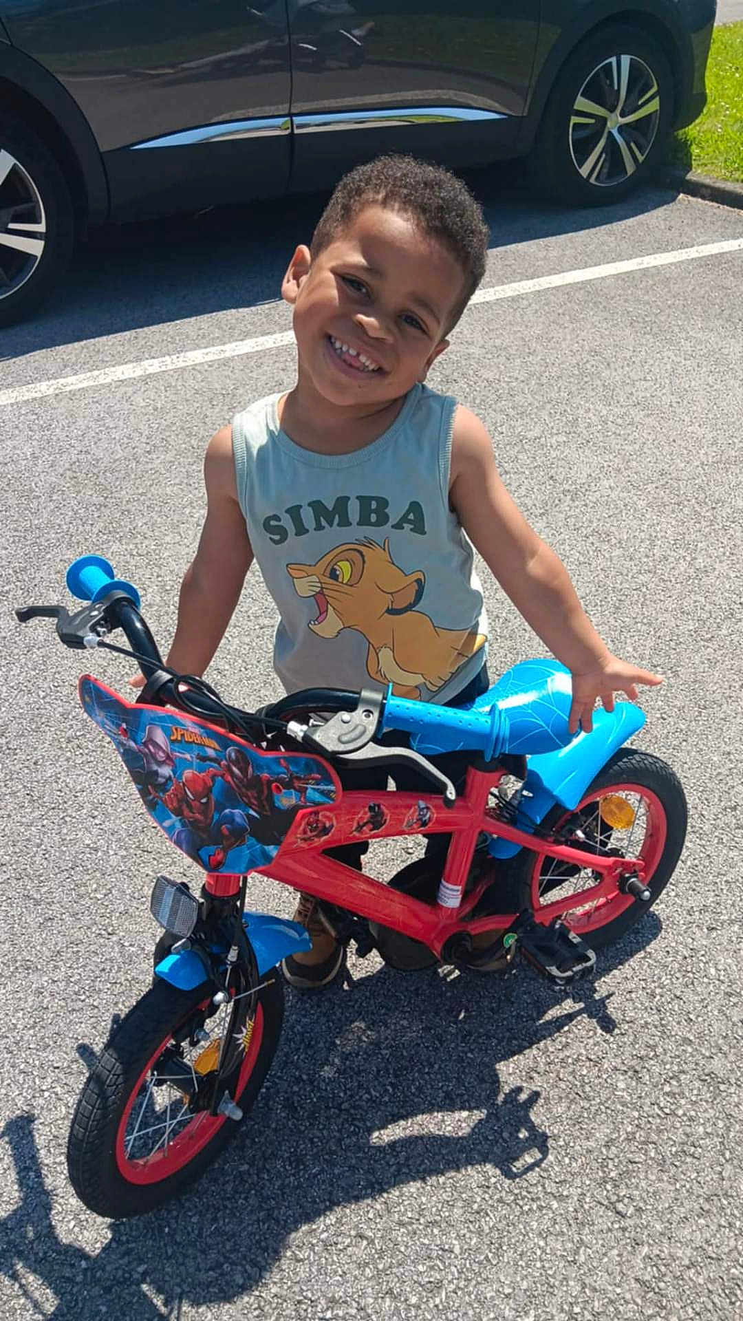 Hector participe au concours pour gagner de l'argent avec cette photo : bicycle, bike, casual_clothing, child, concrete, daylight, handlebar, happy, outdoor, parking_lot, person, shadow, simba, smiling, spiderman, sunny, toy, tshirt, wheel, young