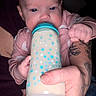 baby, bottle, feeding, infant, hand, tattoo, pink_clothing, blue_stars, milk, closeup, portrait, child, cute, human, skin, fingers, nourishment, drink, person, dark_background