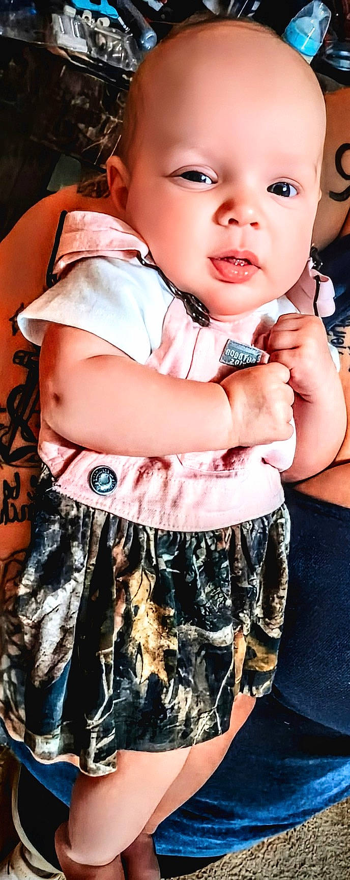 Madison is registered to the contest to win money with this photo: adult, arm, baby, camo_skirt, carpet, child, clothing, cute, expression, face, footwear, hand, indoor, infant, person, pink_clothing, skin, standing, support, tattoo